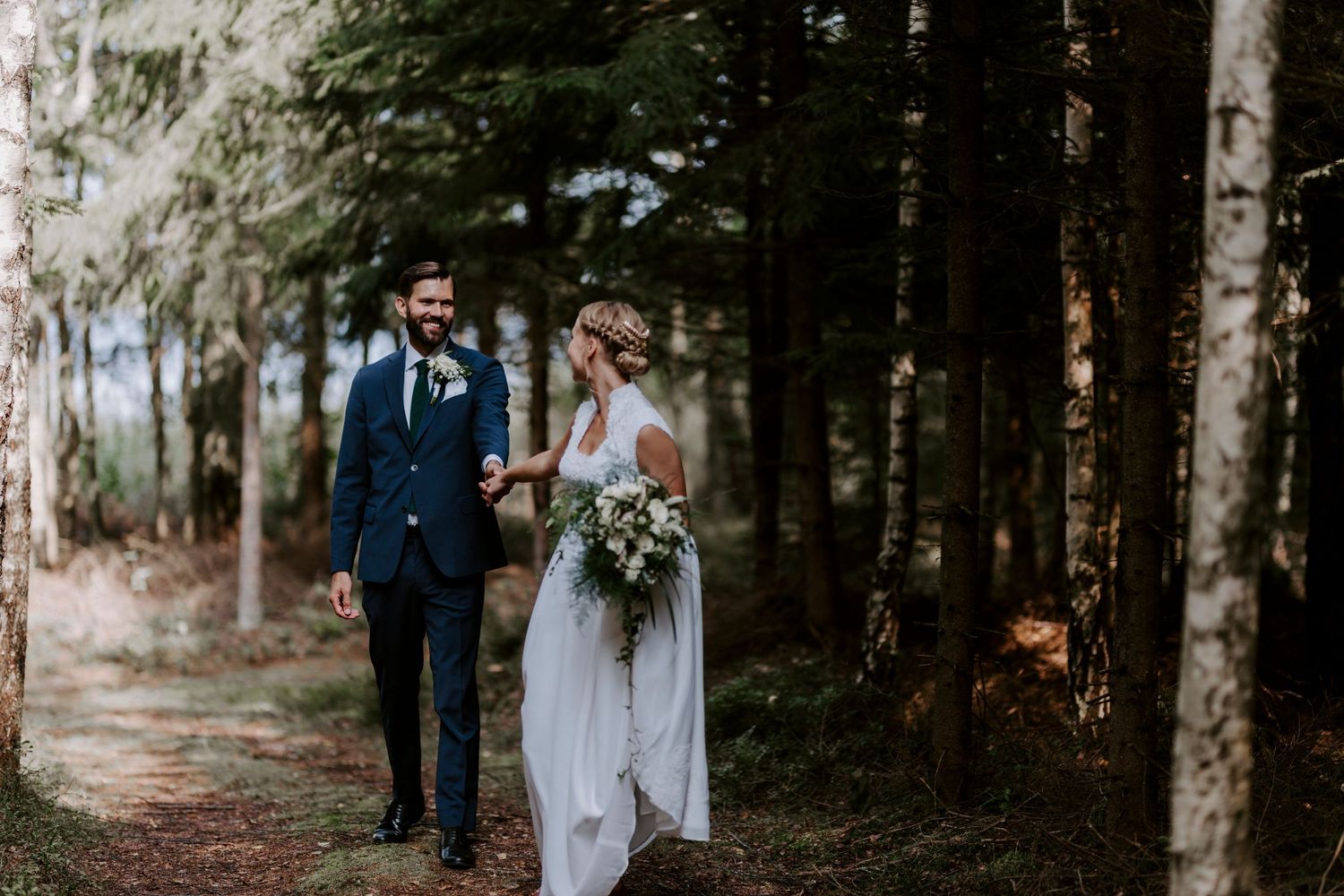 Couple walks hand in hand through forest path in wedding attire.