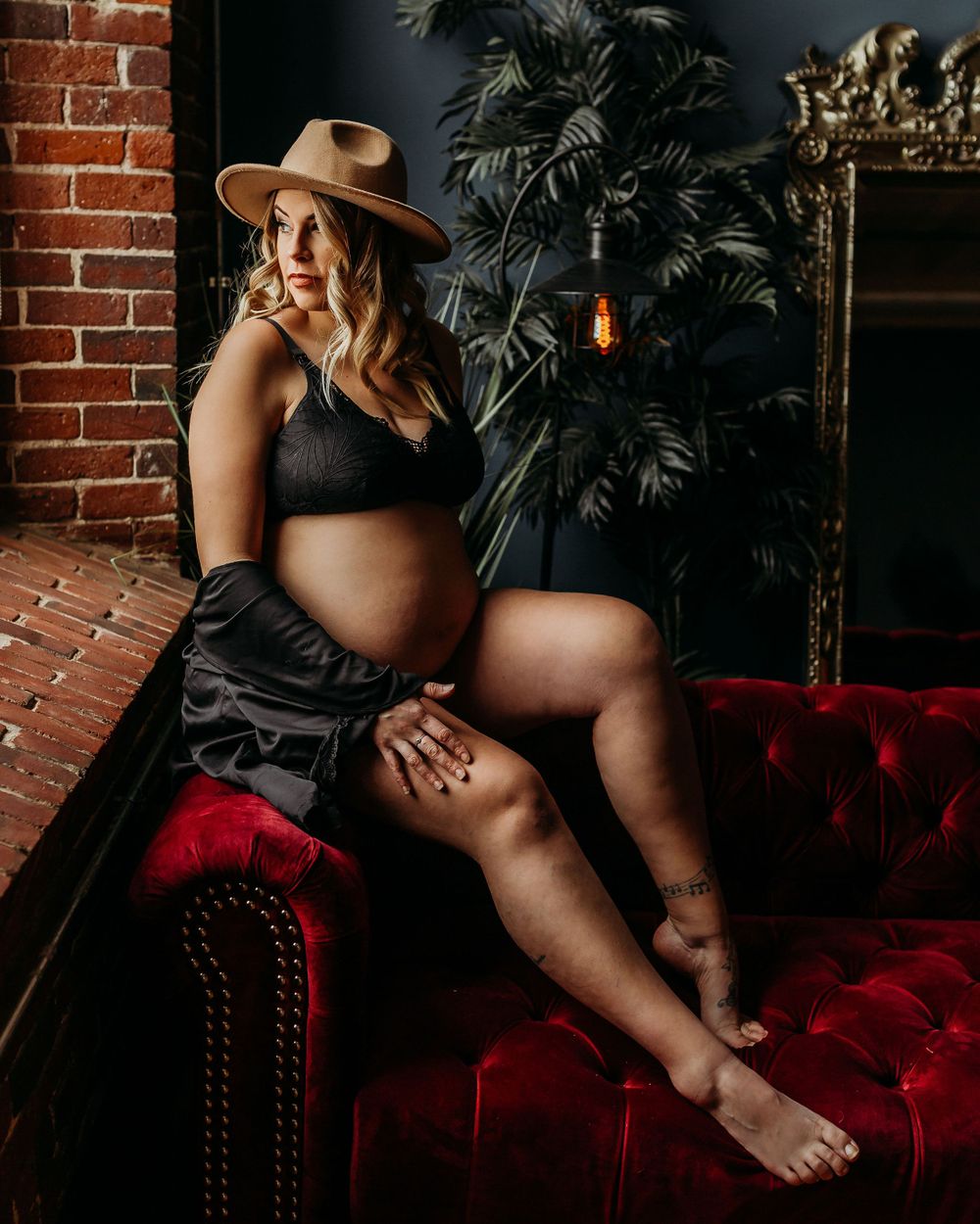 Artistic pregnancy photo with brown fedora hat on burgundy tufted sofa near decorative plants.
