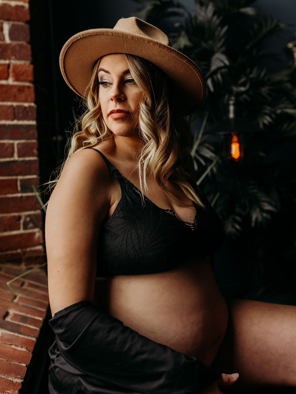 Dramatic maternity portrait with tan hat and black top against industrial brick backdrop.
