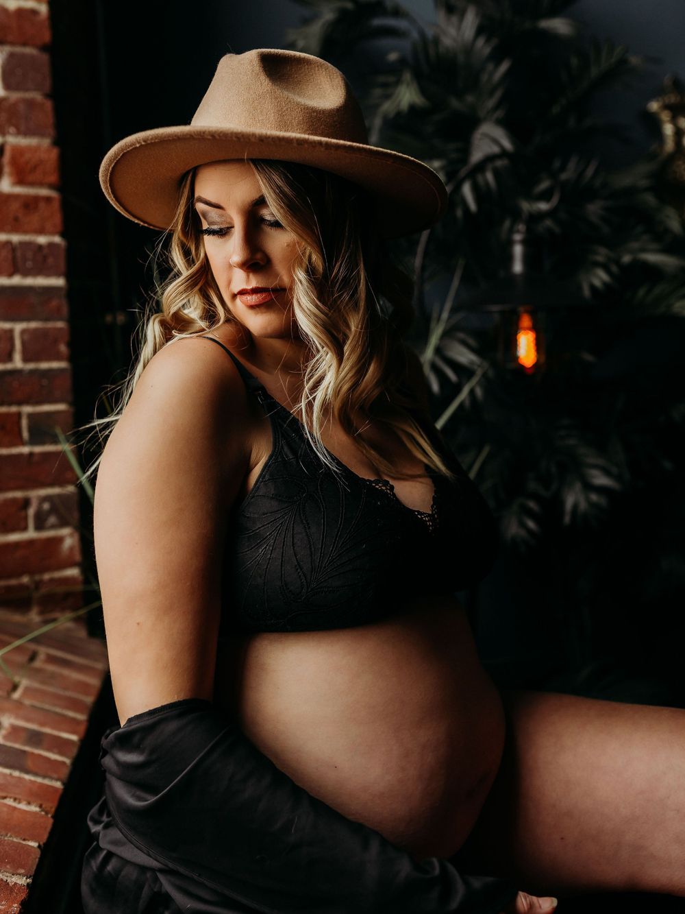 Side-view maternity portrait with brown hat against dark wall with hanging pendant light.