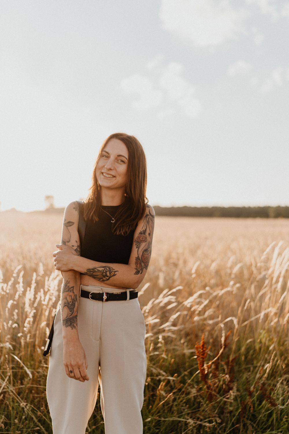 Tattooed woman in black top and light pants standing in golden wheat field at sunset.