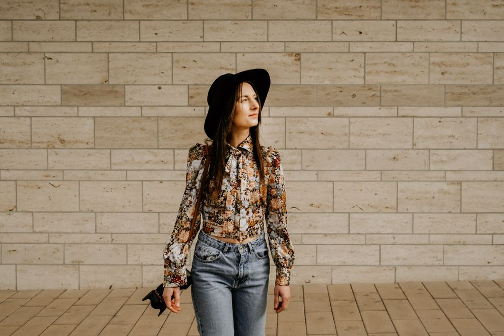 Stylish woman in floral blouse and wide-brim hat against stone wall backdrop.