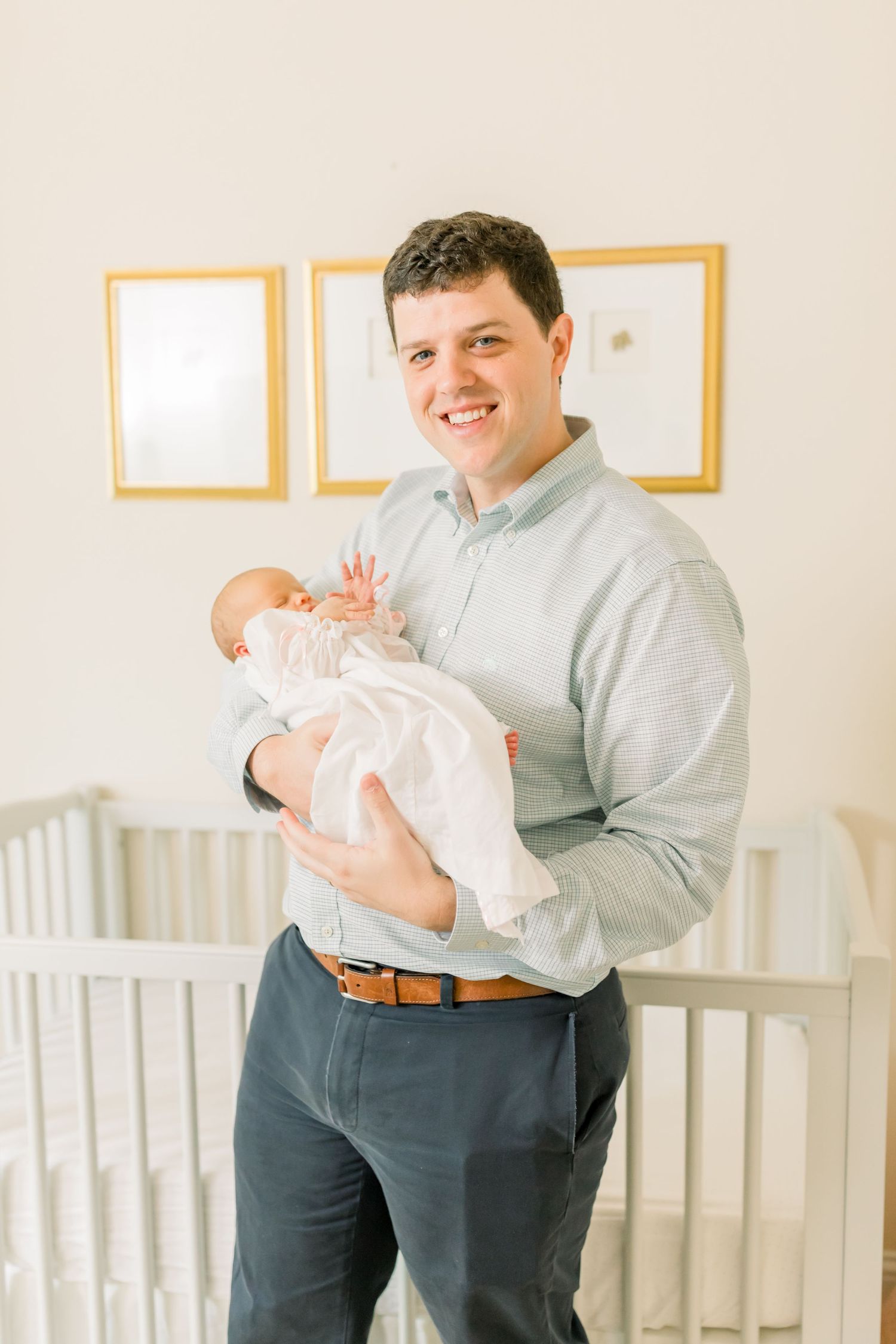 A figure in business casual attire holds a baby in white clothing in front of a nursery crib.