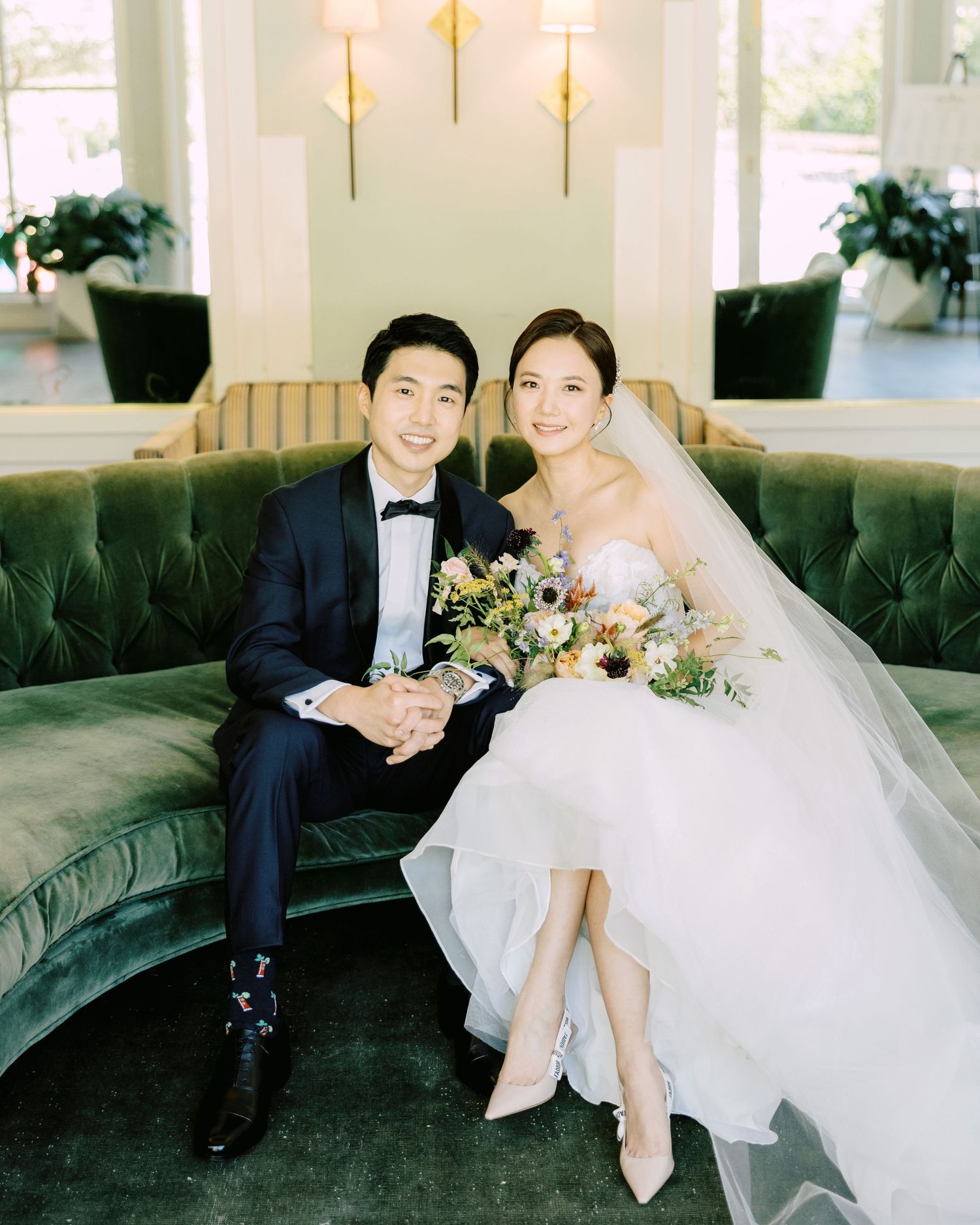 Elegant indoor fine art wedding photography of a bride and groom sitting together on a green velvet sofa.