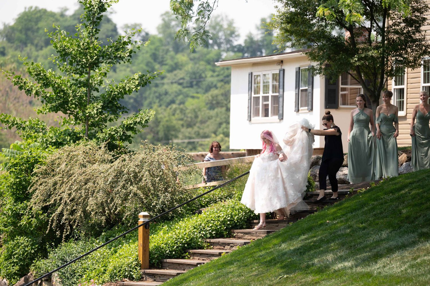 Wedding party descends outdoor stone steps on a hillside near a white house on a sunny day.