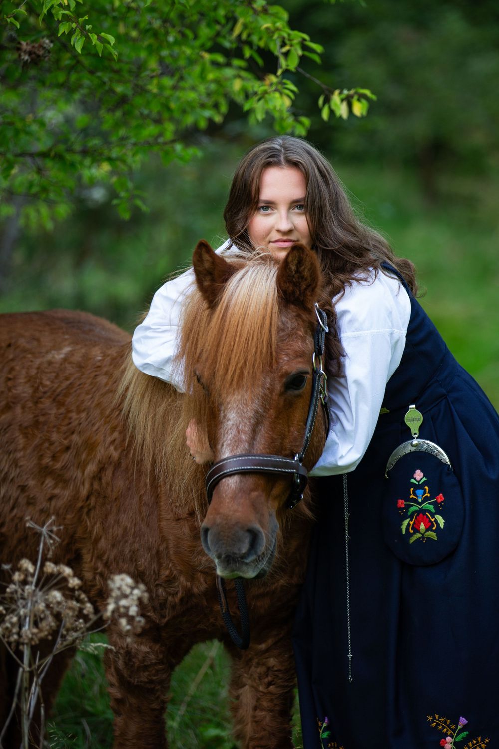 Person in traditional dress embracing brown horse in natural setting.