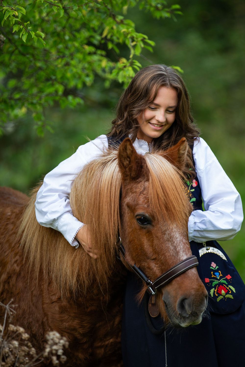 A young person in traditional dress hugs their chestnut brown pony with green foliage in the background.
