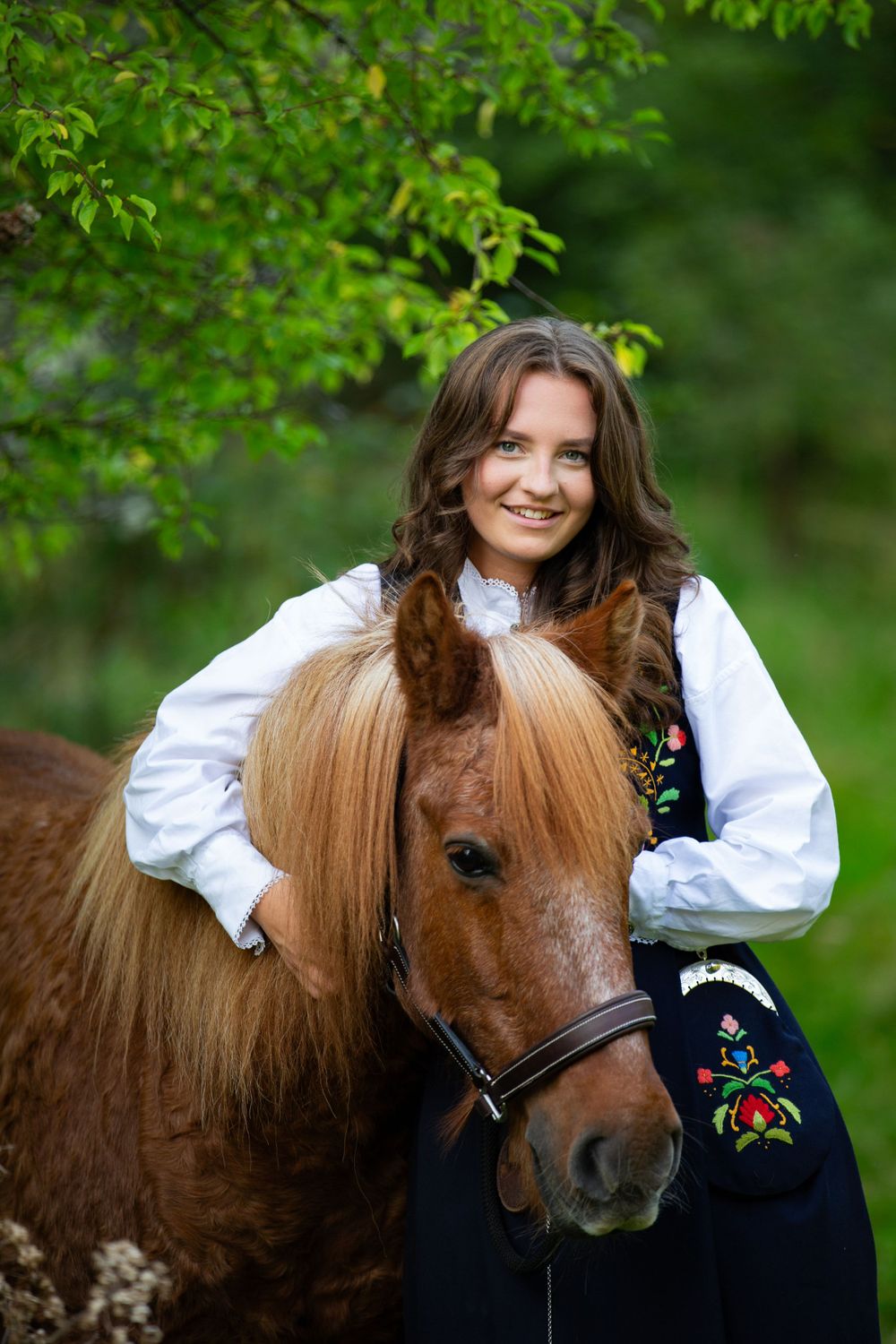 A joyful person in folk dress embraces their brown pony against a leafy green backdrop.
