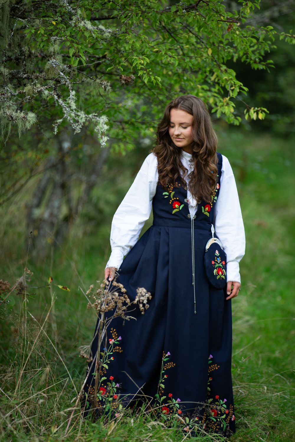Person wearing traditional embroidered dress stands in a lush garden setting with flowering branches.
