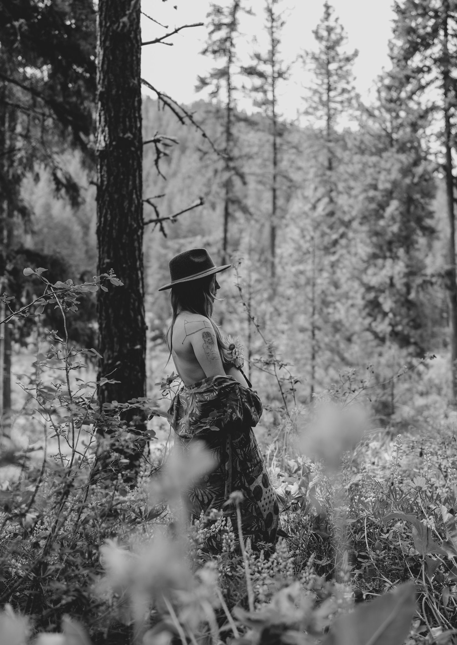 Black and white image of person in hat standing among forest vegetation.