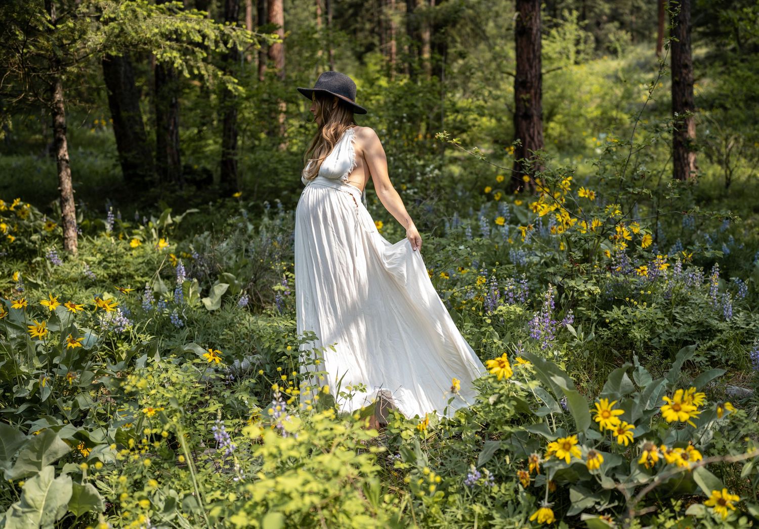 Person in white dress and hat walking through wildflower meadow in forest setting.