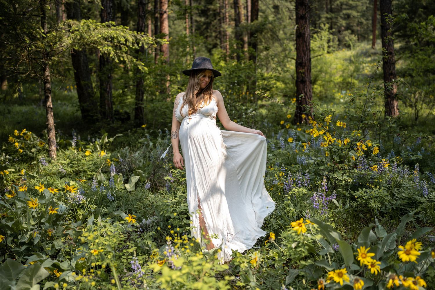 Person in flowing white dress poses in forest meadow filled with yellow flowers.
