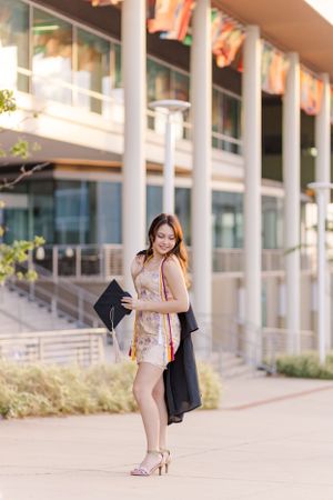 Person in floral dress walking along modern building facade with graduation accessories.