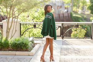 A student wearing a green university sweater and white shorts poses outdoors on a sunny campus walkway.