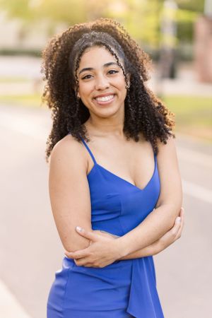 Graduate in blue dress poses against blurred outdoor background.