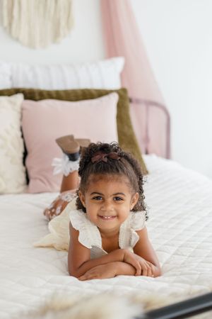 Child lying on white bedding with pink and olive pillows in modern bedroom setting.