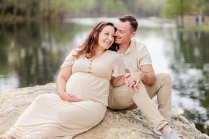 Expecting couple sits on rocky outcrop by peaceful lake during maternity photoshoot.