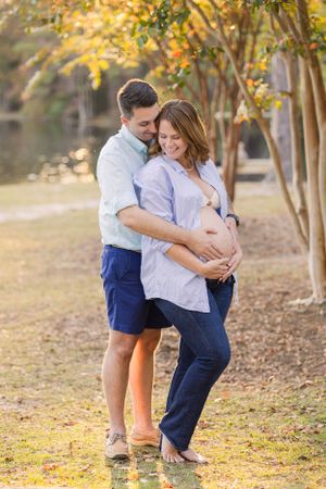 Romantic maternity photo of couple embracing under autumn trees near water.