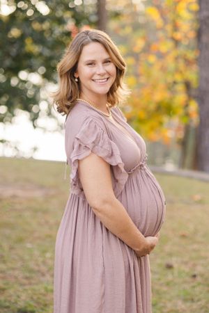 Expectant mother in flowing mauve dress poses outdoors during autumn photoshoot.