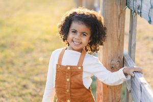 Young girl in brown overalls stands by wooden fence post in golden sunlight.