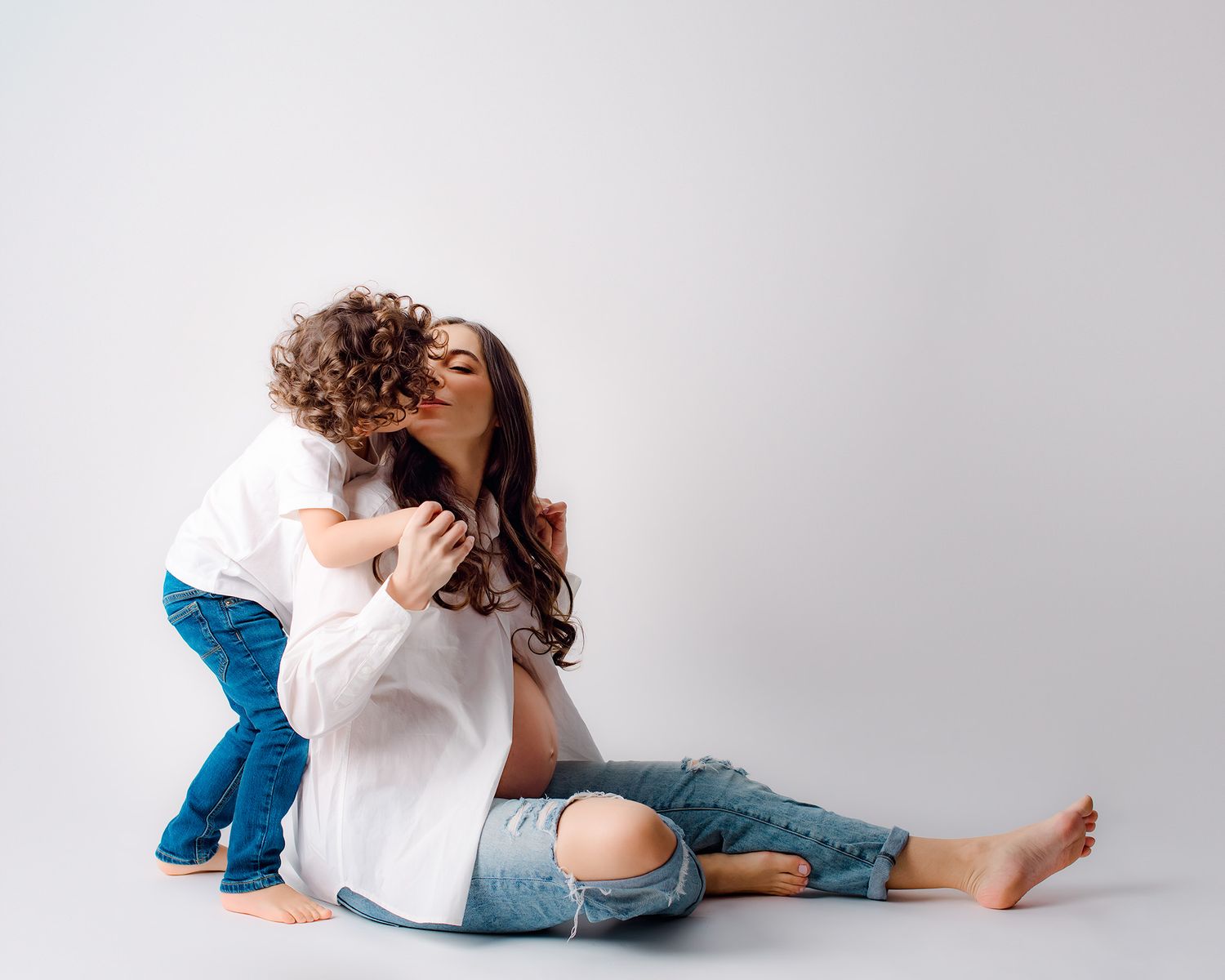 Curly-haired toddler kissing pregnant mother sitting on white studio floor in jeans.