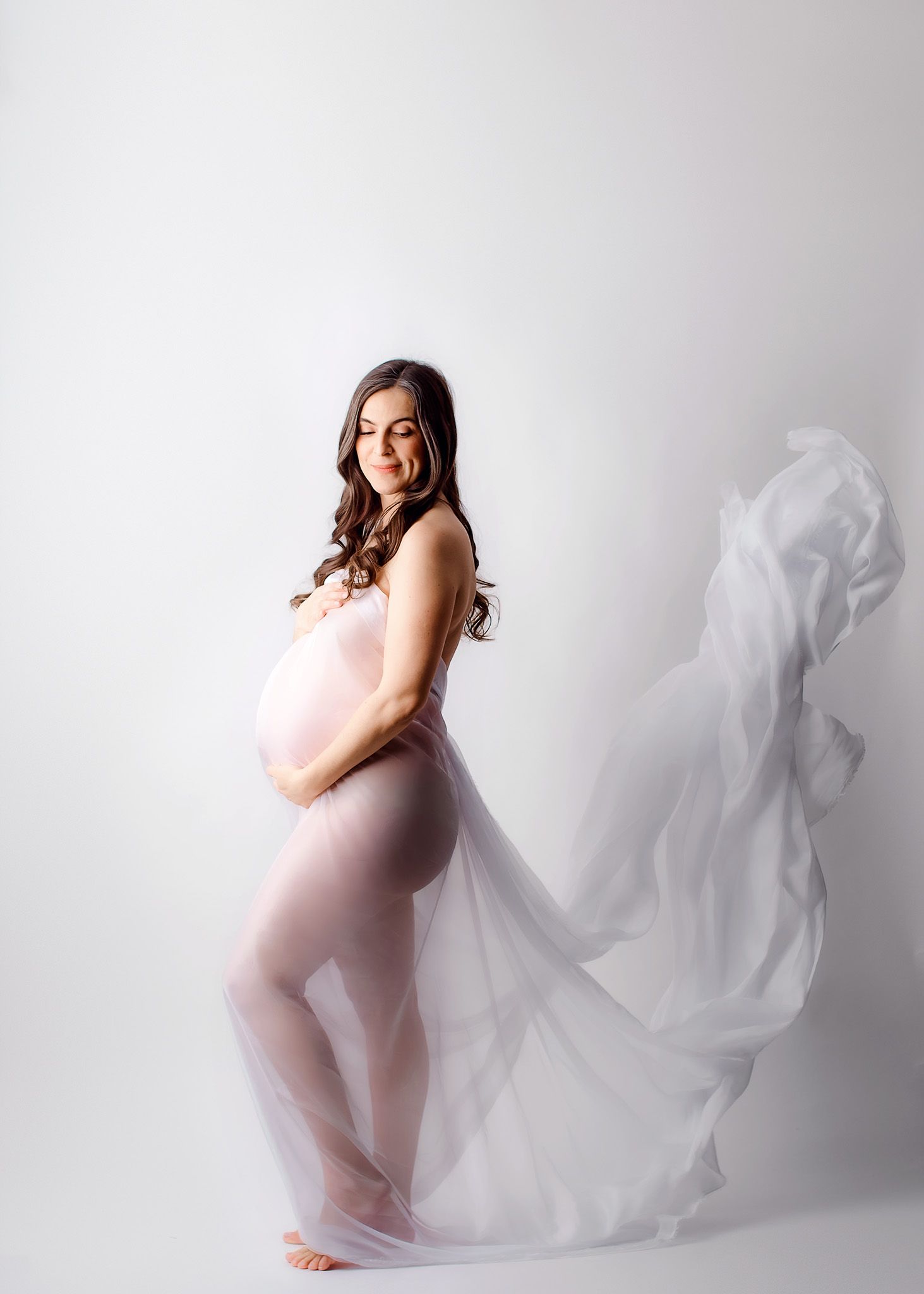 Pregnant woman in flowing white sheer fabric poses gracefully in a studio maternity photoshoot.