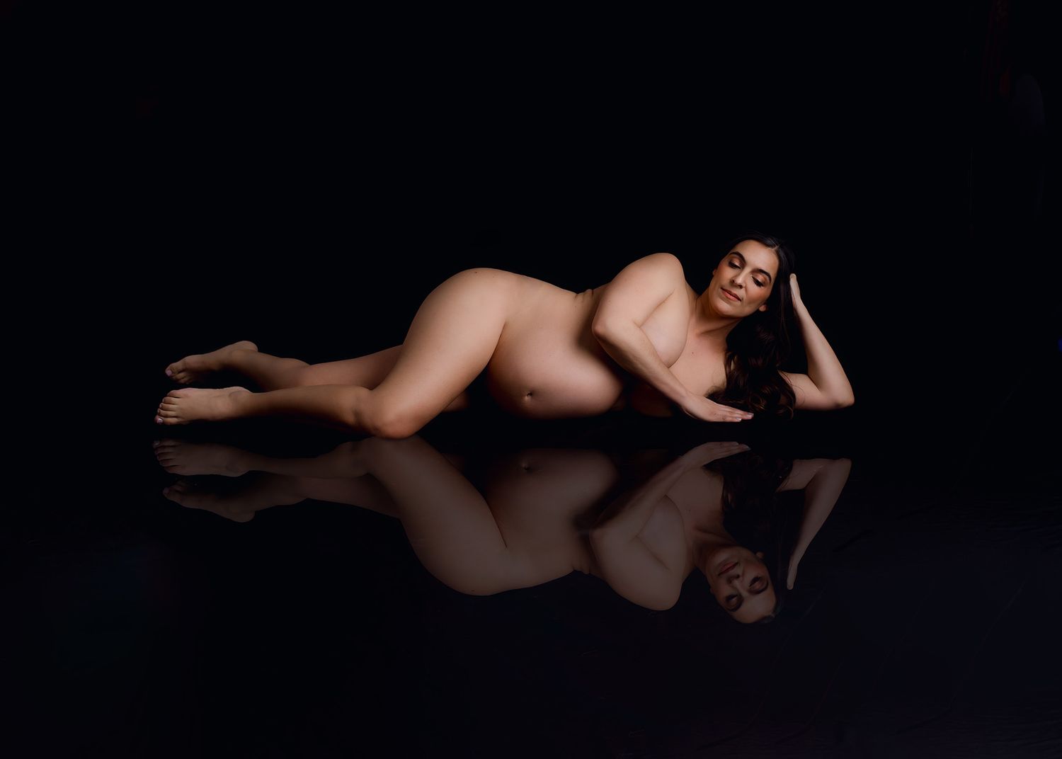 Pregnant woman posed elegantly on reflective black surface in artistic maternity fine art photography studio portrait.