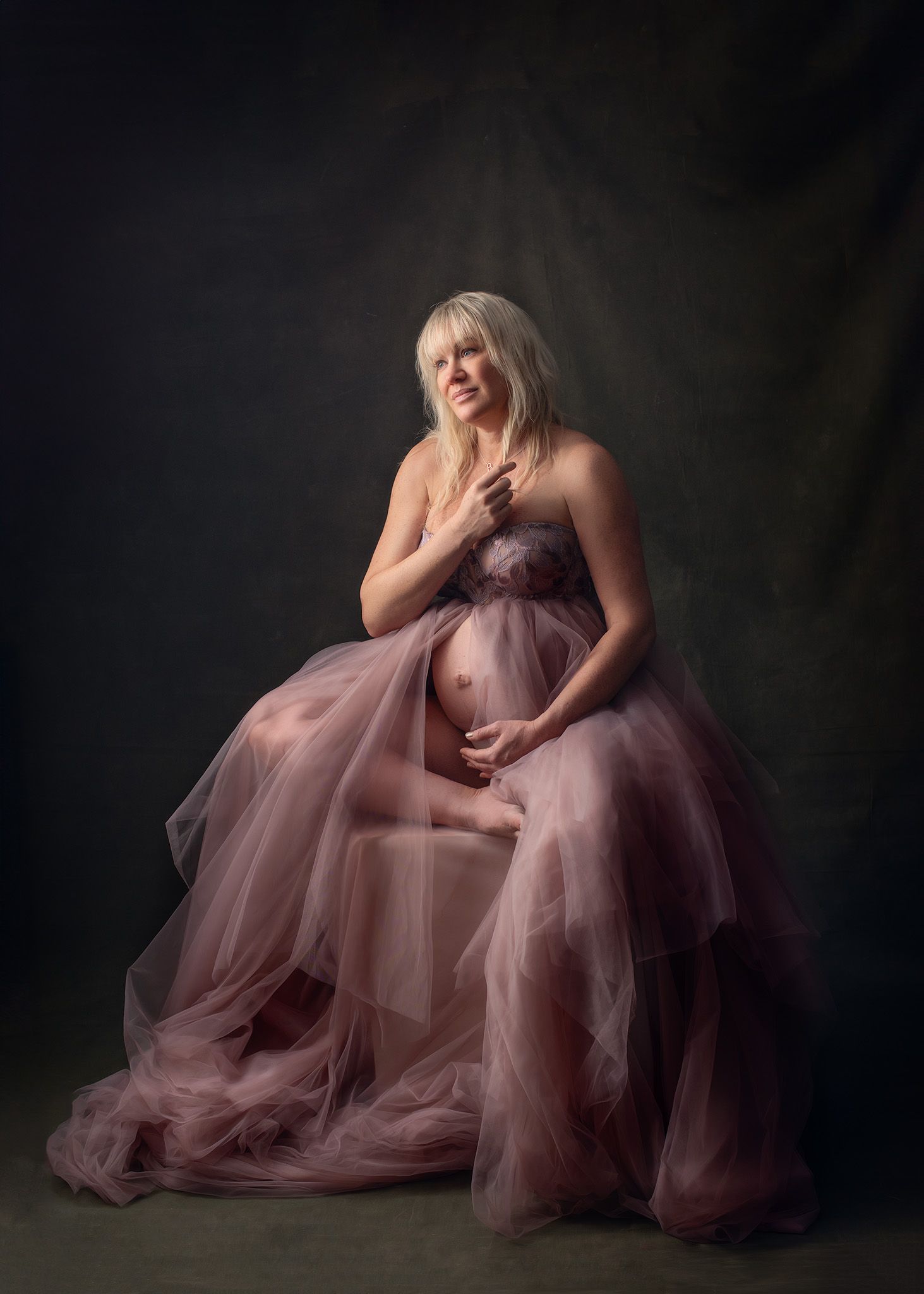 Pregnant woman in a flowing dusty rose tulle gown sitting gracefully in a studio maternity portrait.