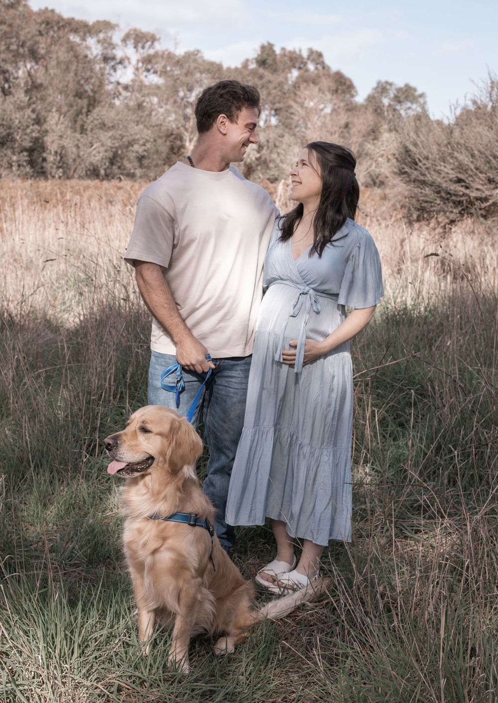 A couple shares a tender moment in a field of tall grass with their golden retriever companion at sunset.