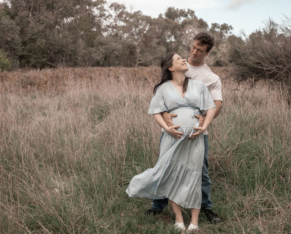 A couple embraces in a rustic field with tall grass during golden hour, wearing soft flowy clothing.
