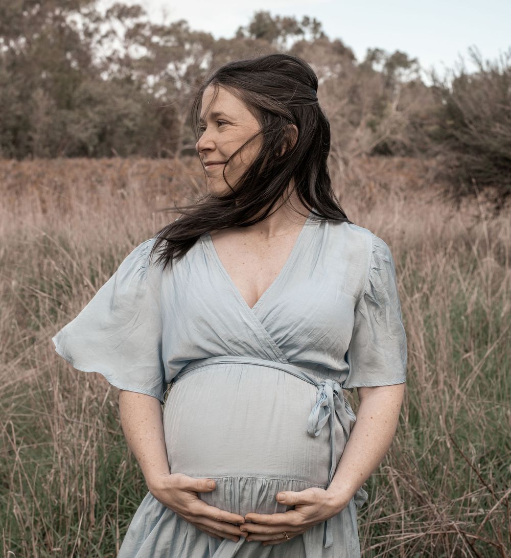 A woman in a flowing light blue dress cradles her baby bump in a serene outdoor maternity photoshoot.