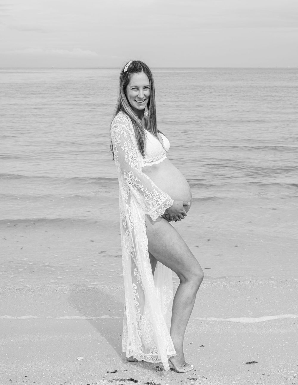 Woman in white lace dress poses playfully on sandy beach during sunset photoshoot.