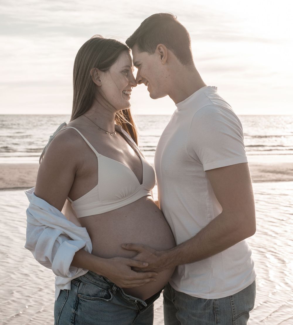 Pregnant couple embracing on beach at sunset, woman in white bralette with exposed belly, man in white t-shirt.
