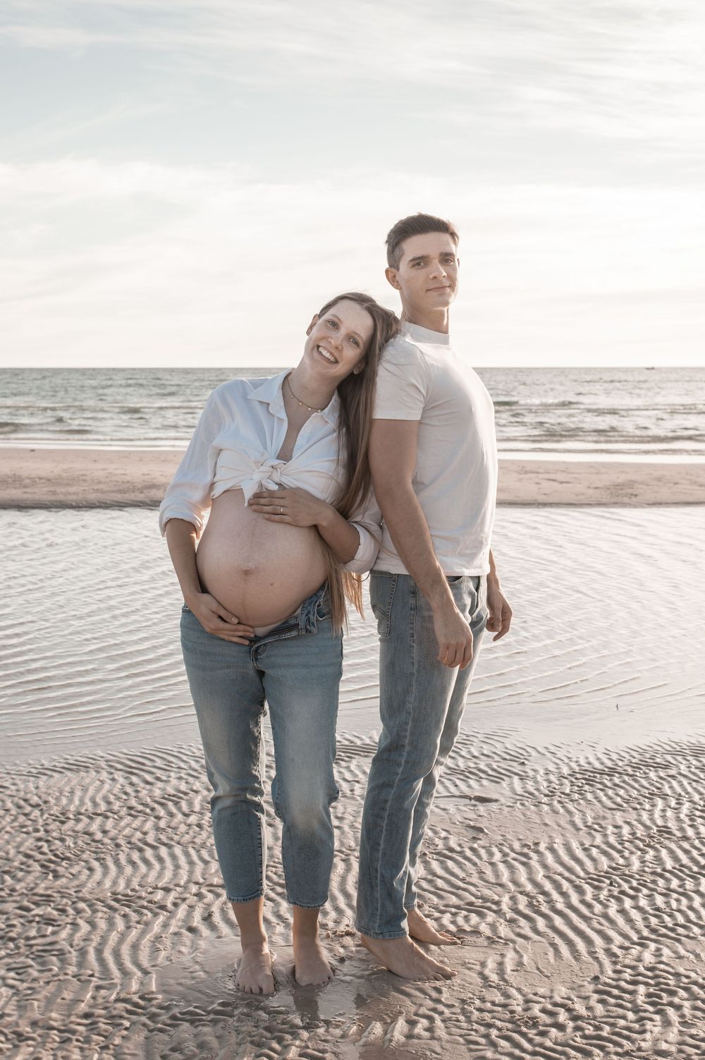 Pregnant couple standing back-to-back on a sandy beach, woman cradling her baby bump, both smiling in casual attire.
