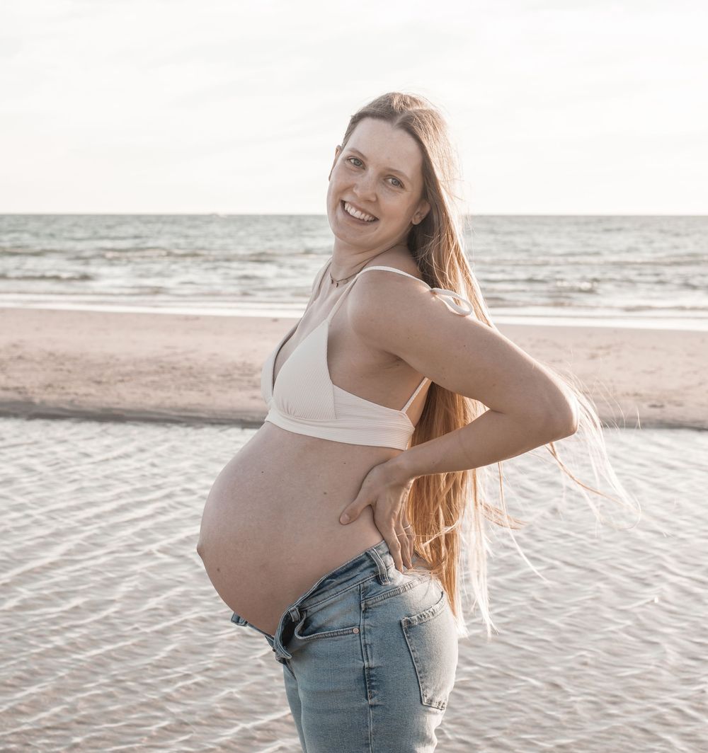 Smiling pregnant woman in white crop top and jeans posing on beach with ocean backdrop.