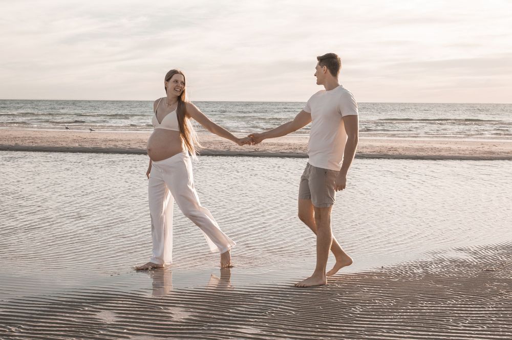 Pregnant woman and man holding hands on a serene beach at sunset, wearing white outfits, barefoot on wet sand.