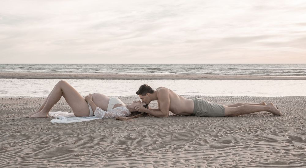 Pregnant couple sharing a tender kiss while lying on a sandy beach at sunset.