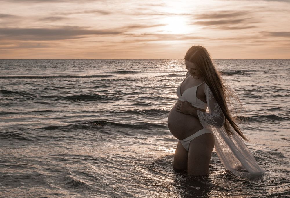 Pregnant woman in white bikini standing in ocean waves at sunset with flowing fabric, maternity beach photography.