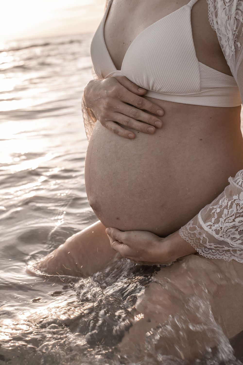 Pregnant woman in white bikini top cradling her baby bump while standing in shallow ocean water at sunset.