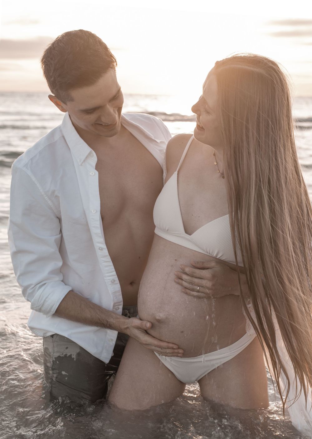 Couple at beach sunset, pregnant woman in white bikini with partner touching her baby bump in shallow ocean water.