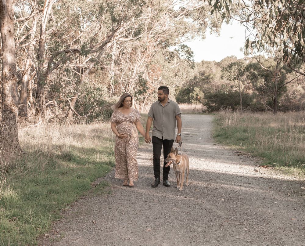 Pregnant couple walking a dog on a scenic tree-lined path during a maternity photo shoot outdoors.