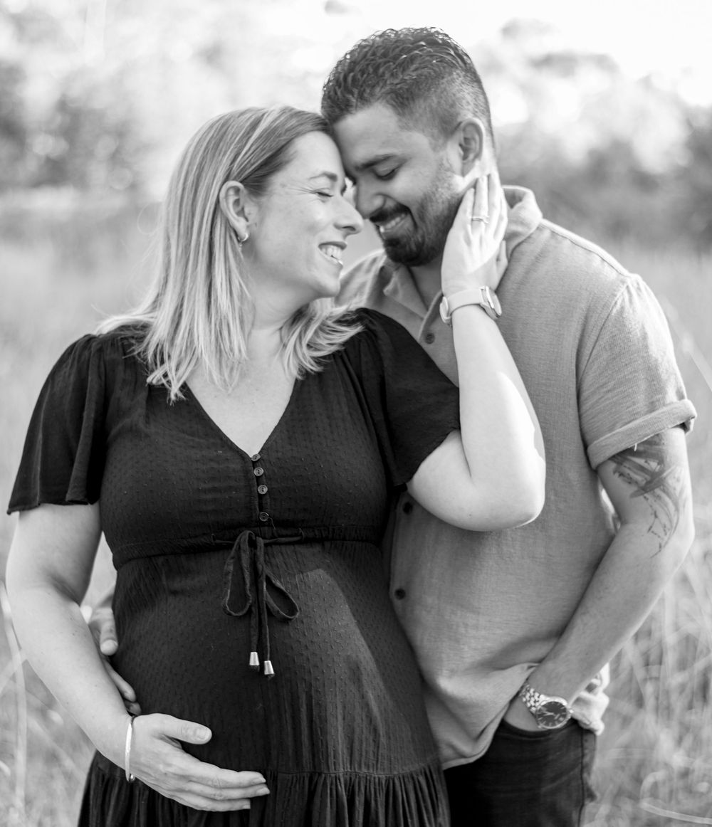 Black and white photo of a smiling pregnant couple sharing a tender moment outdoors in a field.