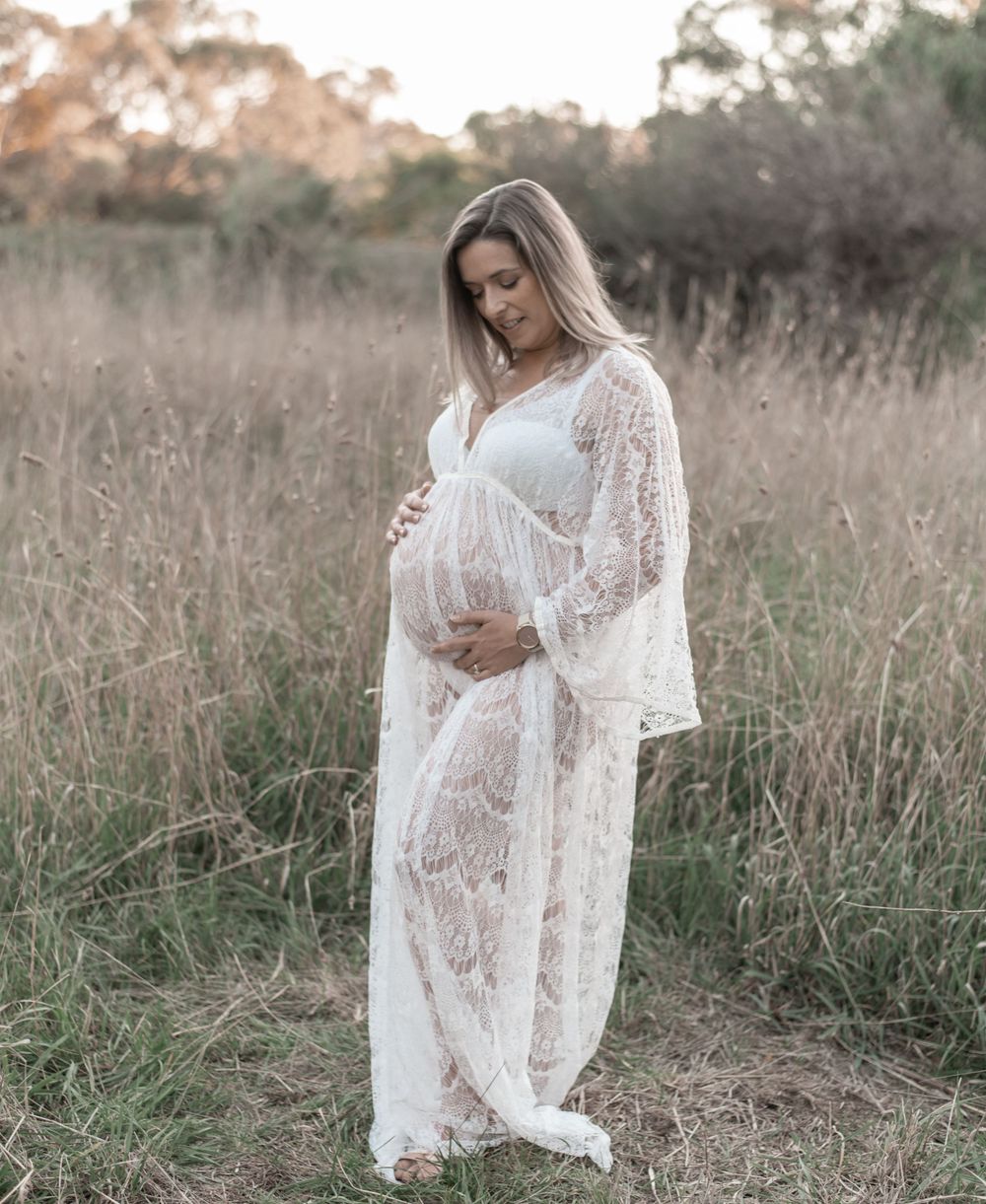 Pregnant woman in white lace maternity gown standing in golden field at sunset, cradling baby bump.