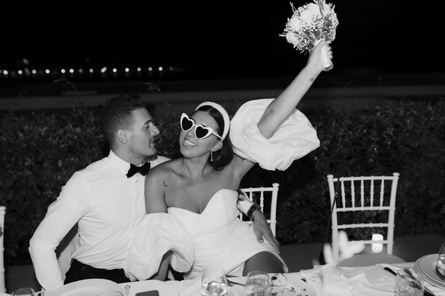 A stylish couple in black and white celebrating at a wedding reception table with one person raising their bouquet.