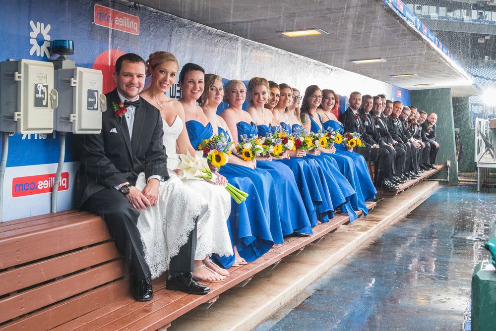 Wedding party in formal attire seated on dugout bench with blue and black color scheme in Citizens Bank Park.