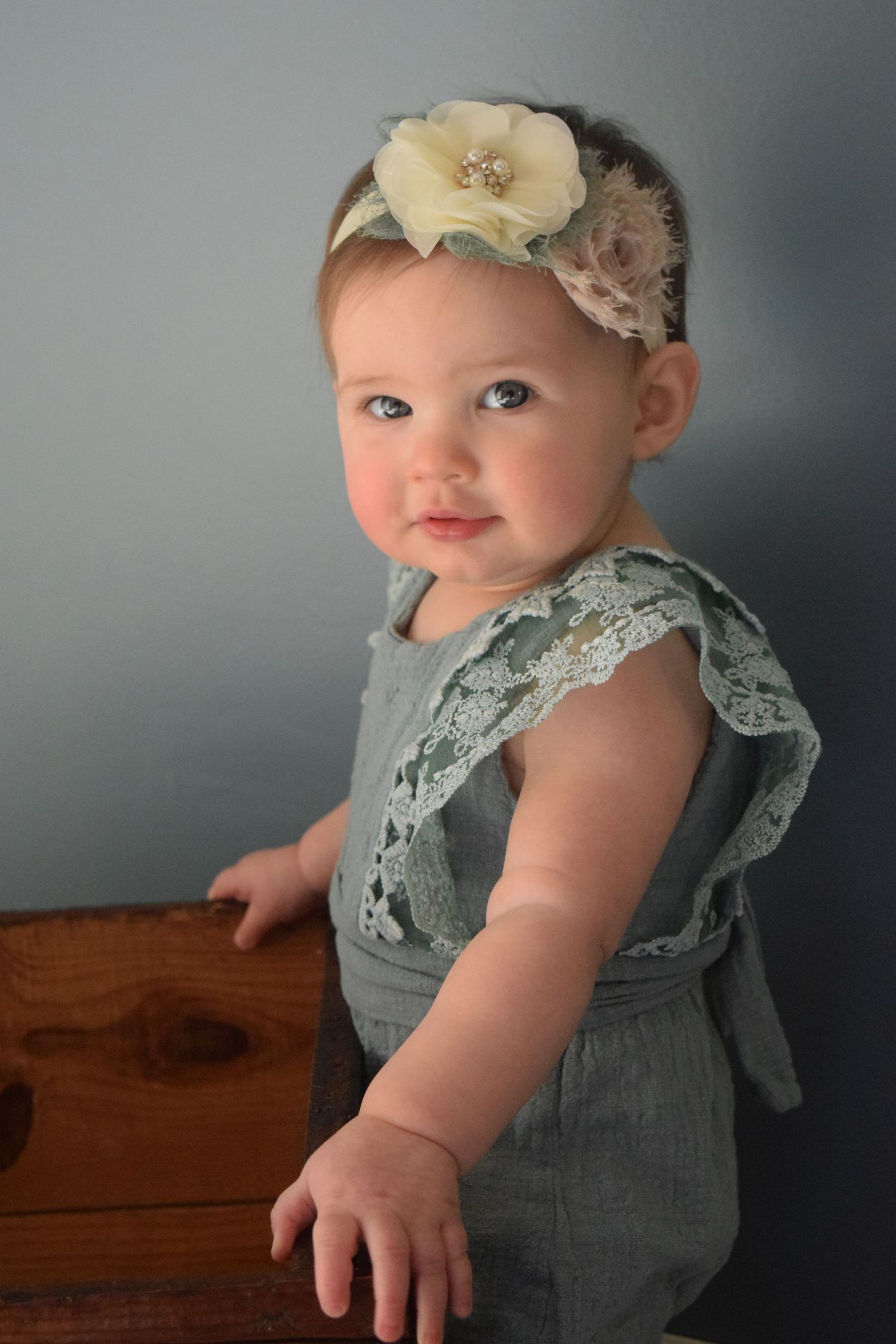 Baby in sage green lace romper poses against gray backdrop wearing cream floral headband.