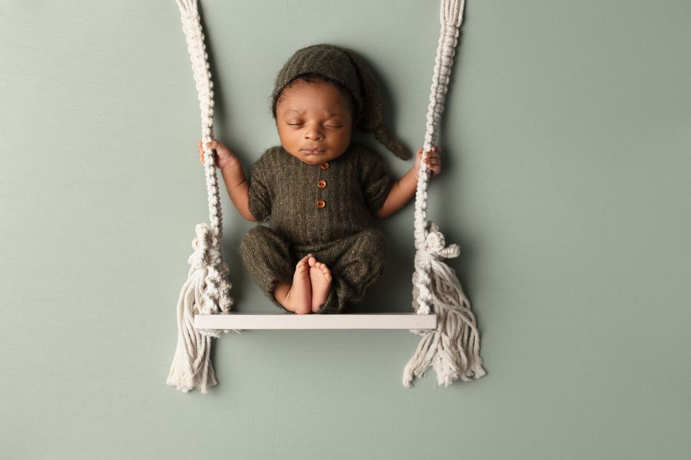 Baby wearing knit sweater sitting in macrame swing against gray wall.