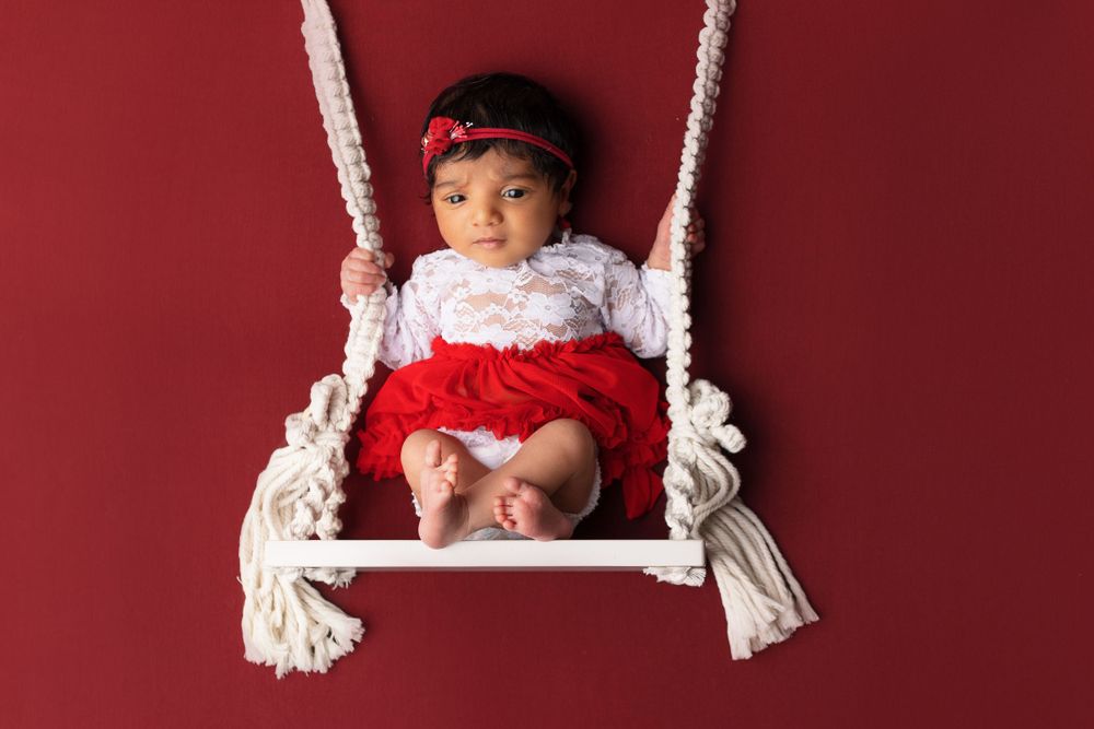 Newborn on white rope swing against red backdrop wearing white and red outfit.