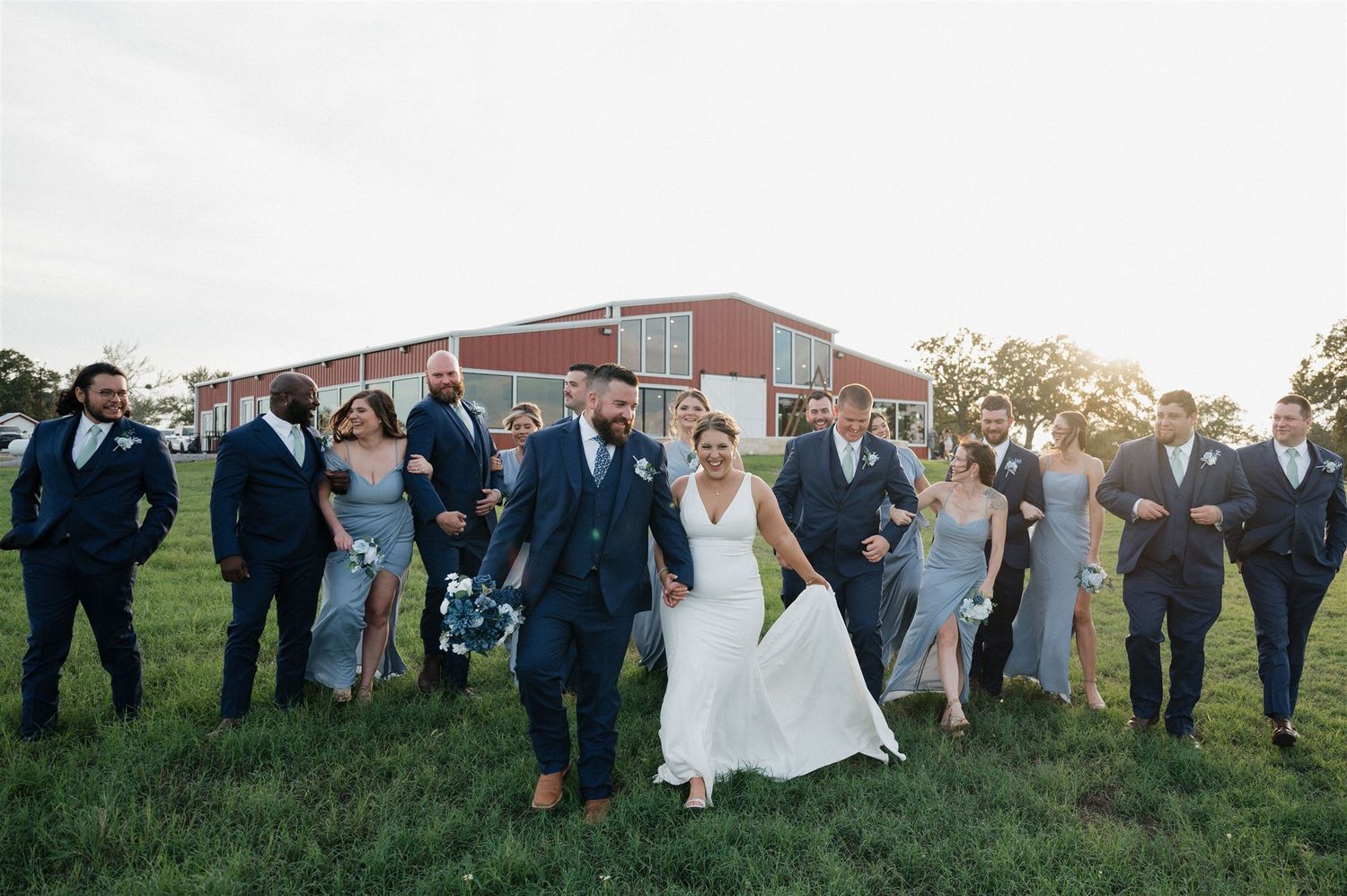 Wedding party poses together on grassy field during sunset celebration.