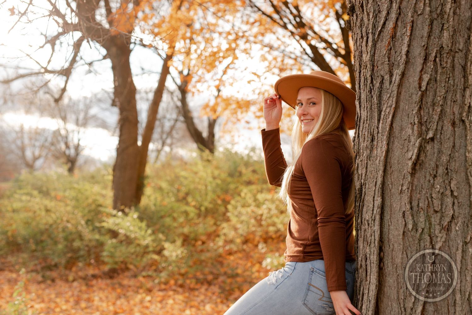 Person in autumn attire poses beside tree trunk with golden fall leaves in background.
